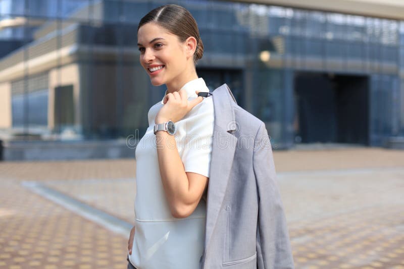 Situación sonriente de la mujer de negocios con una chaqueta sobre su hombro cerca del edificio de oficinas imagen de archivo Situación sonriente de la mujer de negocios con una chaqueta sobre su hombro cerca del edificio de oficinas imagen de archivo