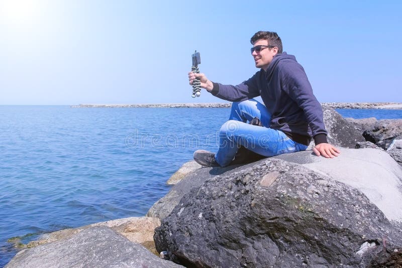 Sitzt Videoschwätzchen touristischen Smartphone Gespräch des Mannes in Meer mit kleinem Stativ auf Stein stockfotografie
