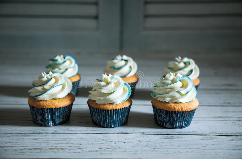 Close-up of six cupcakes with white and blue cream on top against white wooden background