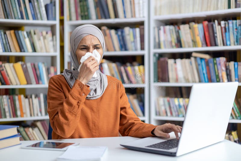 A Muslim woman in a hijab sneezes into a tissue while working at a laptop in a public library, depicting illness and persistence in education and work environments. A Muslim woman in a hijab sneezes into a tissue while working at a laptop in a public library, depicting illness and persistence in education and work environments.