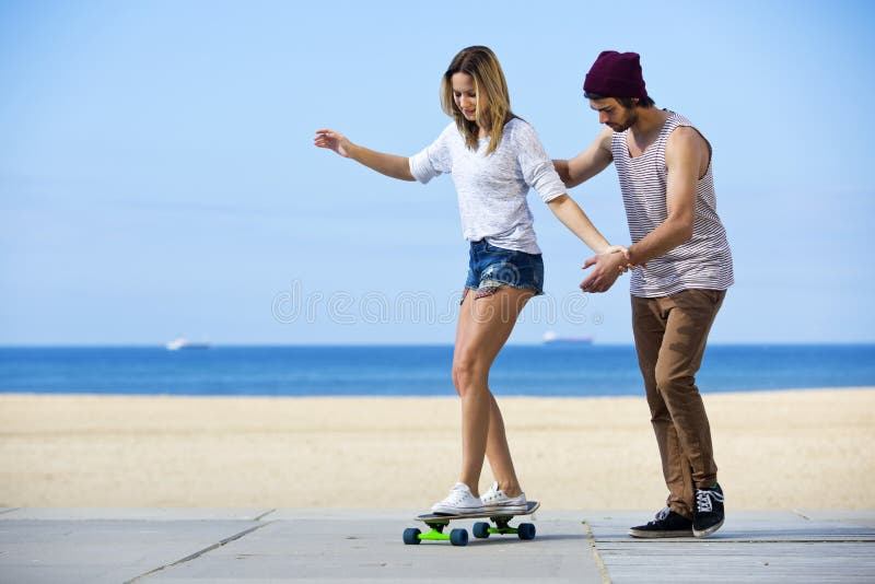 Young men gives his girlfriend her first skateboarding lesson on a seaside boulevard, during a lovely, calm summer afternoon. Young men gives his girlfriend her first skateboarding lesson on a seaside boulevard, during a lovely, calm summer afternoon.