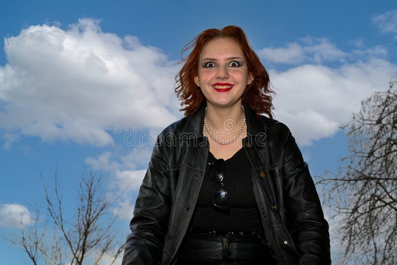 Young woman dressed in black, with a smiling face and wide eyes, stands against a blue sky, photographed from the bottom up. Young woman dressed in black, with a smiling face and wide eyes, stands against a blue sky, photographed from the bottom up