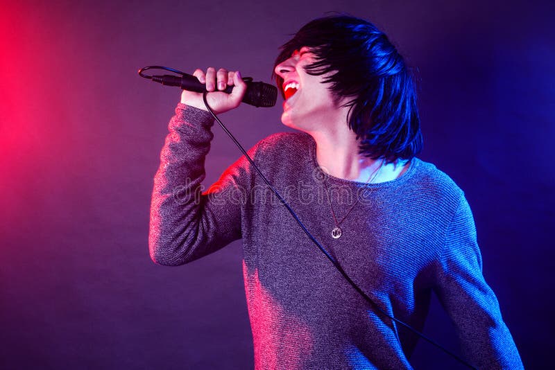 The young guy vocalist is screaming in microphone on stage on concert illuminated with red and blue lights. The young guy vocalist is screaming in microphone on stage on concert illuminated with red and blue lights