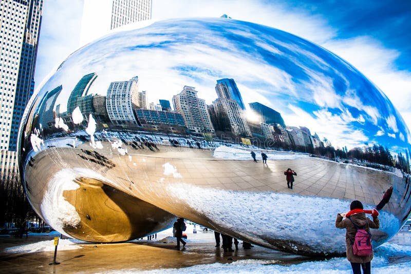Skygate Bean covering by snow against high building towers and blue sky with unidentified visitors at Millenium Park.