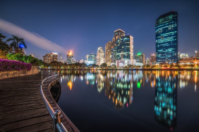 Skyscrapers and Lake with Wooden Walk Way in City Park. View of Benjakiti Park at Night. Beautiful Night Scene of Public Park in Bangkok, Thailand, Asia. Skyscrapers and Lake with Wooden Walk Way in City Park. View of Benjakiti Park at Night. Beautiful Night Scene of Public Park in Bangkok, Thailand, Asia