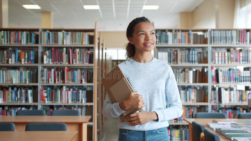 Skyvlig tonårsflicka som går med böcker i skolbiblioteket och ler