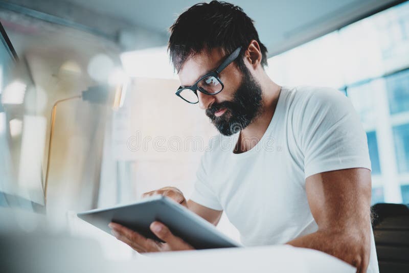 Skäggig man, i exponeringsglas och att använda för öga för vit tshirt bärande den bärbara elektroniska pro-minnestavladatoren på royaltyfri bild Skäggig man, i exponeringsglas och att använda för öga för vit tshirt bärande den bärbara elektroniska pro-minnestavladatoren på royaltyfri bild