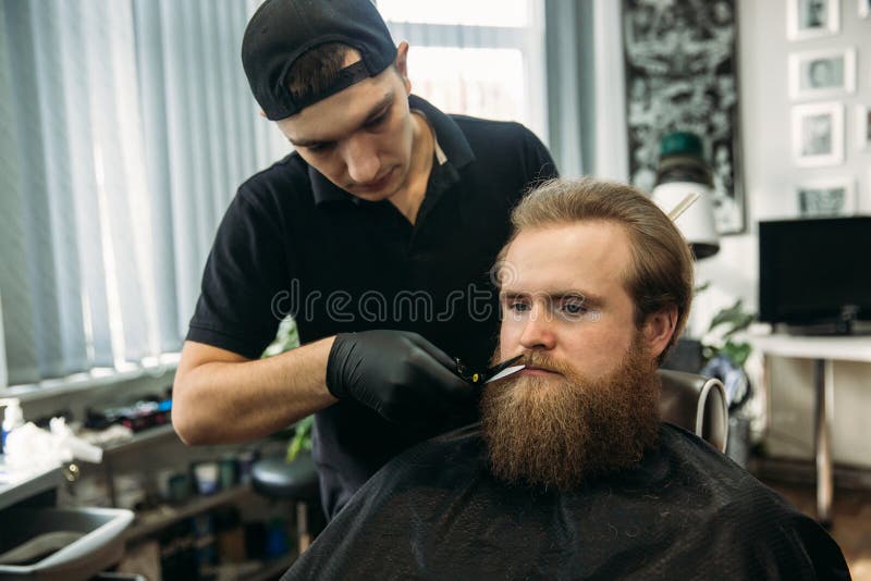 Skäggig man med det långa skägget som får stilfullt hår som rakar, frisyr, med rakkniven av barberaren i frisersalong royaltyfria bilder Skäggig man med det långa skägget som får stilfullt hår som rakar, frisyr, med rakkniven av barberaren i frisersalong royaltyfria bilder