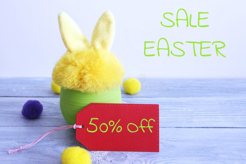 Yellow fur toy bunny in a green plastic pot on gray wooden boards, and on a gray background with the words Easter sale, a red paper tag with the words `50% off` and yellow and purple wool balls. Yellow fur toy bunny in a green plastic pot on gray wooden boards, and on a gray background with the words Easter sale, a red paper tag with the words `50% off` and yellow and purple wool balls