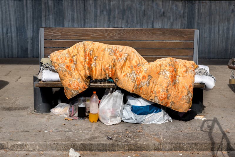 Slechte dakloze mens of vluchtelingsslaap op de houten bank op de stedelijke die straat in de stad met een deken met zakken van k royalty-vrije stock afbeelding Slechte dakloze mens of vluchtelingsslaap op de houten bank op de stedelijke die straat in de stad met een deken met zakken van k royalty-vrije stock afbeelding