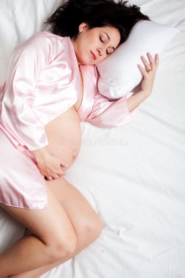 Beautiful Caucasian pregnant brunette woman in pink satin robe night gown peacefully sleeping in a bed with white sheets while holding her tummy belly. Beautiful Caucasian pregnant brunette woman in pink satin robe night gown peacefully sleeping in a bed with white sheets while holding her tummy belly.