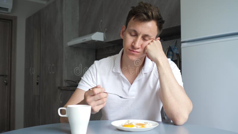 Sleepy man has breakfast and drinks coffee in kitchen