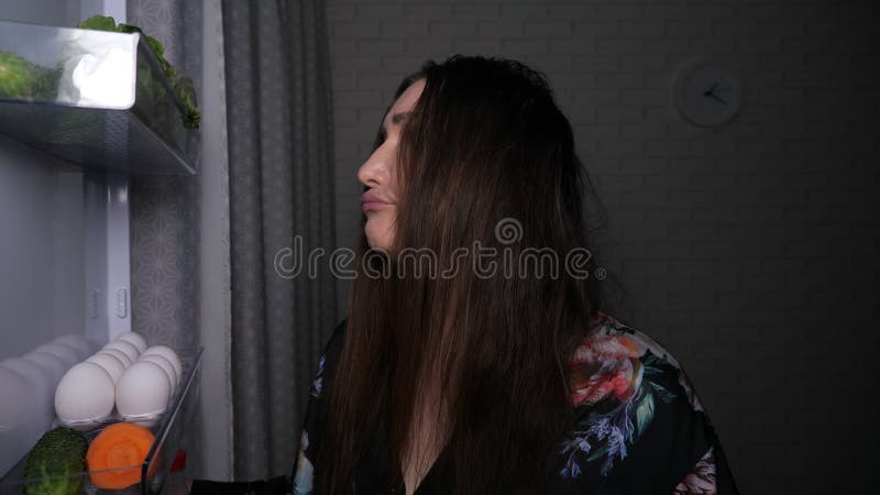 Sleepy woman takes fresh carrot to eat from refrigerator