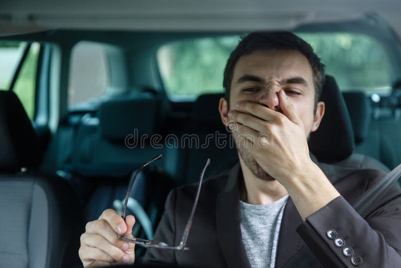 Sleepy young guy yawns while covering his mouth with his left hand. He is holding glasses at his right hand. Road safety concept