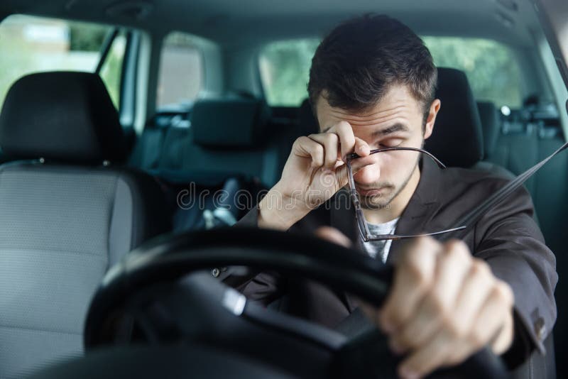 Sleepy young man rubs his eyes with his right hand. His left hand is on the steering wheel. He is sitting at his car. Road safety