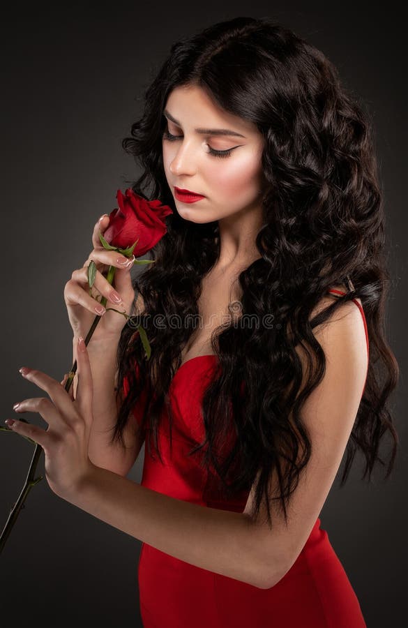 A slender brunette woman with long curly hair and beautiful makeup, she is wearing a red dress, she is holding a rose