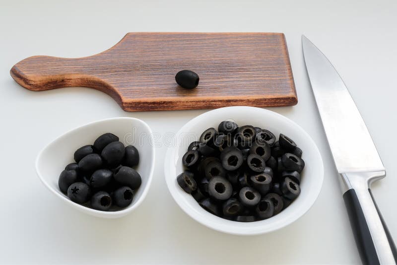 Sliced olives and whole black olives in a white bowls, brown wooden cutting board and sharp chef knife on a kitchen table.