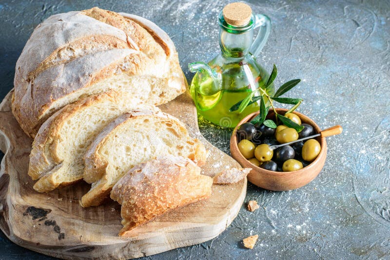 Slices of white homemade bread on the wooden cutting board. Olive oil bottle and olives in the wooden bowl on the dark background