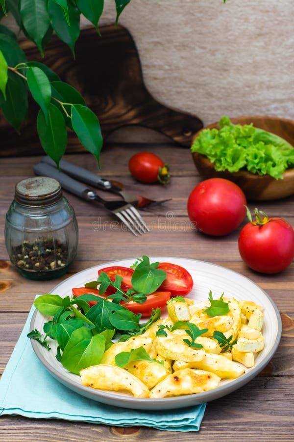 Slices of zucchini baked in spices with cheese on a plate with tomatoes and arugula on a wooden table royalty free stock photo Slices of zucchini baked in spices with cheese on a plate with tomatoes and arugula on a wooden table royalty free stock photo