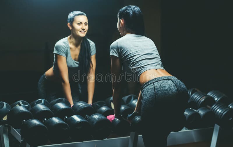 Slim, bodybuilder girl, lifts heavy dumbbell standing in front of the mirror while training in the gym. stock photo Slim, bodybuilder girl, lifts heavy dumbbell standing in front of the mirror while training in the gym. stock photo