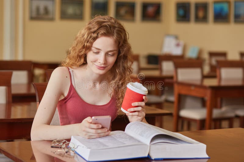 Slim good looking young student sitting in local library, holding papercup of coffee and smartphone, looking at mobile screen