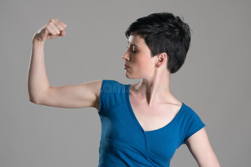 Slim young short hair woman flexing arm bicep muscle over gray studio background.
