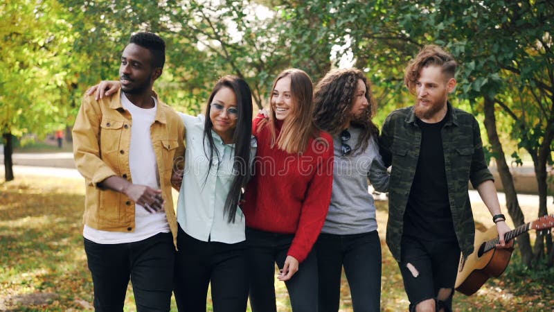 Slow motion of happy young people friends walking together in park in autumn holding guitar talking and laughing. Youth