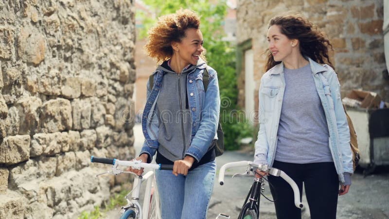 Slowmotion счастливых кавказских и Афро-американских туристов девушек смеясь над и идя с велосипедами вдоль улицы с