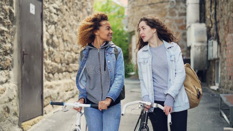 Slowmotion счастливых привлекательных туристов молодых женщин смеясь над и идя с велосипедами вдоль улицы с красивая старой