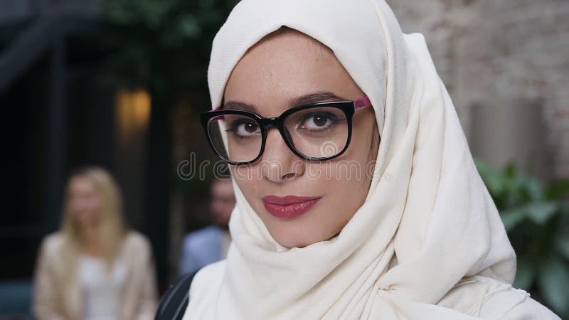 Sluit het portret van een aantrekkelijke moslimvrouw in een bril met een witte hoofddoek op een hijab en kijk naar de camera en g