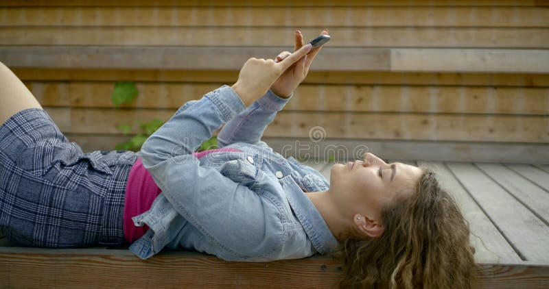 Close-up of curly young girl lying on a wooden bridge on her back and looking at the phone. Close-up of curly young girl lying on a wooden bridge on her back and looking at the phone.