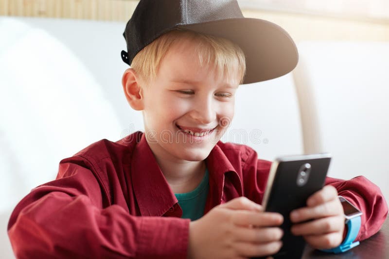 Slut upp av den lyckliga le pojken som bär det svarta locket och röda skjortan som surfar internet på hans mobiltelefon som ser s royaltyfri foto Slut upp av den lyckliga le pojken som bär det svarta locket och röda skjortan som surfar internet på hans mobiltelefon som ser s royaltyfri foto