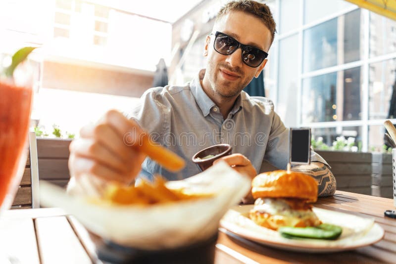 Slut upp av den unga attraktiva mannen som äter den fransmansmåfiskar och hamburgaren på gatakafét royaltyfri bild Slut upp av den unga attraktiva mannen som äter den fransmansmåfiskar och hamburgaren på gatakafét royaltyfri bild