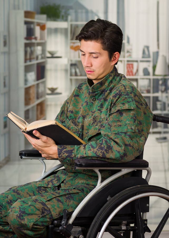 Slutet av stiligt barn tjäna som soldat upp sammanträde på hjulstol som hemma läser en bok, i en suddig bakgrund arkivbilder Slutet av stiligt barn tjäna som soldat upp sammanträde på hjulstol som hemma läser en bok, i en suddig bakgrund arkivbilder