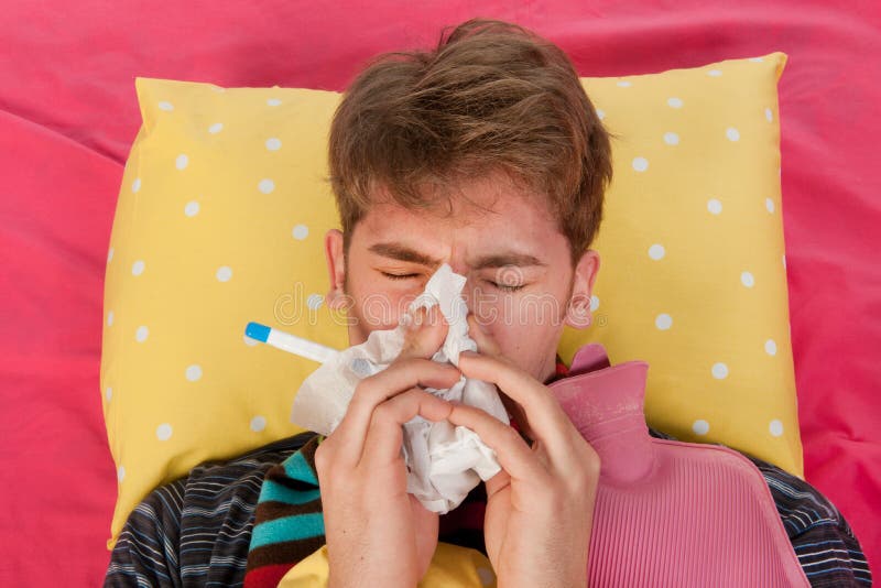 Very ill man islaying in his bed with warm water bottle and thermometer while blowing his nose. Very ill man islaying in his bed with warm water bottle and thermometer while blowing his nose