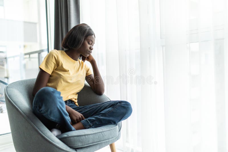 Smaklig, stressad afrikansk flicka som sitter i stolen och sitter hemma och tillbringar helgen ensam royaltyfri fotografi