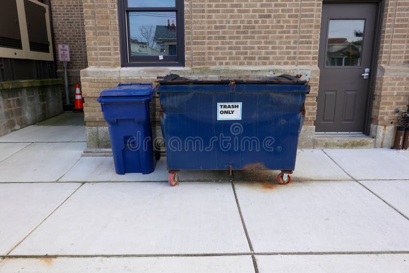 Small Blue Dented Dumpster On Wheels On A Sidewalk Next To A Blue Trash