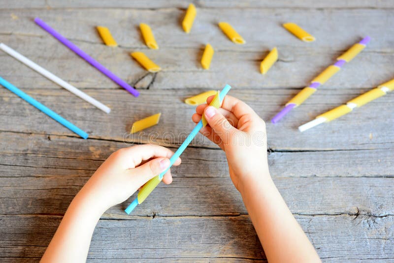 Small child holds straw and dried tube pasta in his hands. Child stringing pasta onto straw