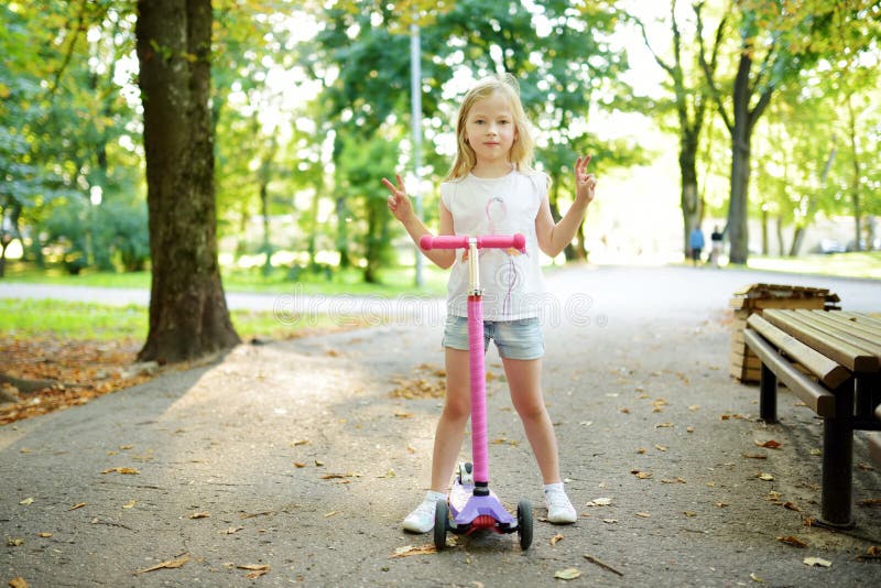 Small child learning to ride a scooter in a city park on sunny summer evening. Cute little girl riding a roller