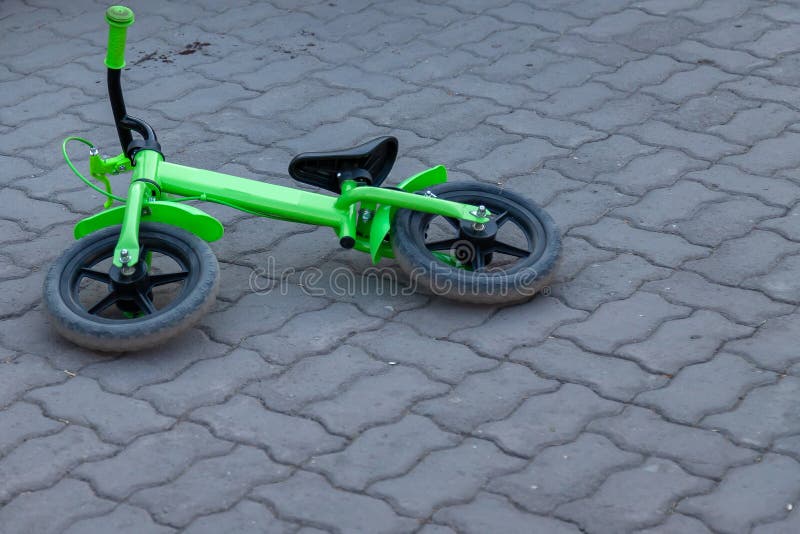 A small childrenâ€™s two-wheeled bicycle of green color lies on its side on a street tile of gray color without people and nobody