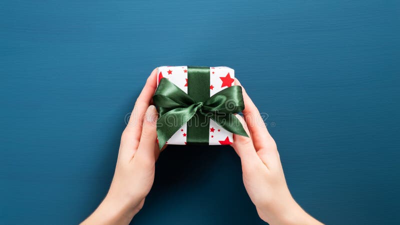 Small Christmas present with green ribbon bow in woman hands. Female hands holding gift box wrapped festive white paper over dark