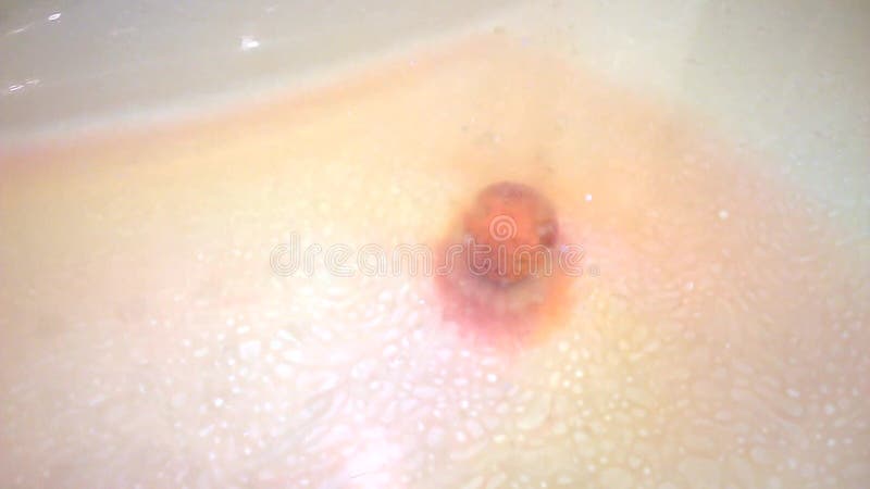 Small colorful bath bomb lies on the bottom in the tub and slowly dissolves. On the surface of the water traces of aromatic oil.