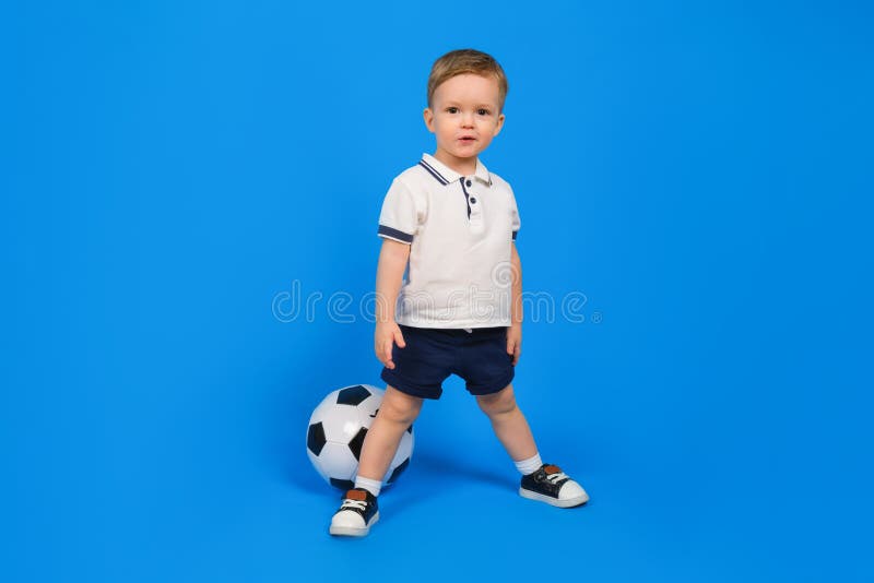 Small happy boy in football uniform and a soccer ball stands on a blue background. football champion. stock photography Small happy boy in football uniform and a soccer ball stands on a blue background. football champion. stock photography