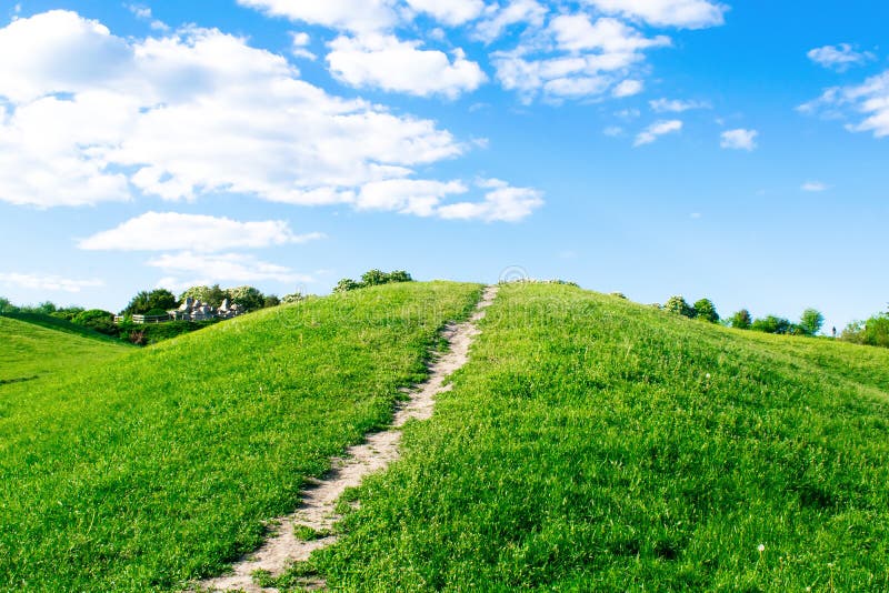 Small hiking trail, path climbing up the mountain, hill, mound with green grass against bright blue sky with clouds