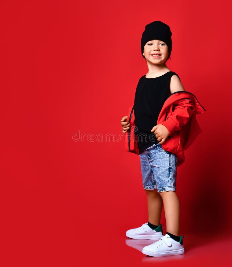 Small positive boy in denim shorts, red jacket, cap white sneakers standing, smiling and looking aside over red wall background