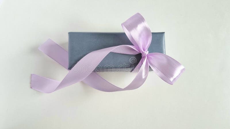 A small rectangular box wrapped in gray paper rests on a white background. It features a glossy, light purple satin ribbon tied into a bow on top. The ribbon's ends trail gracefully beside the box, creating an elegant appearance. Rea trail stock images, royalty-free photos and pictures