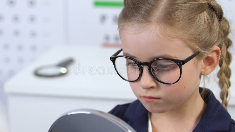 Small schoolgirl in eyeglasses looking in mirror unhappy with appearance, optics