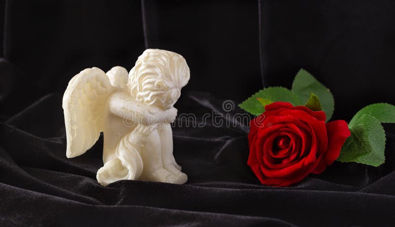 A small sculpture of a baby angel with wings and a red rose on a black background. The concept of funerals, condolences, and