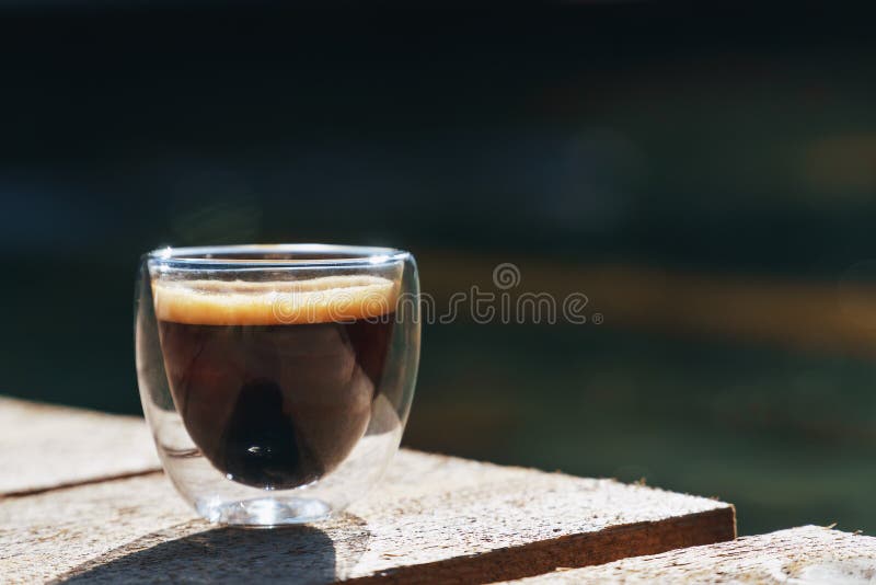 Small transparent double wall glass cup of fresh brewed coffee placed on a natural rough wooden surface in front of a decorative stock image Small transparent double wall glass cup of fresh brewed coffee placed on a natural rough wooden surface in front of a decorative stock image