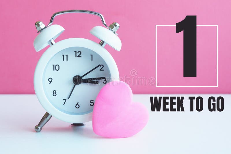 A small white alarm clock and a pink heart figurine. The inscription `1 week to go.
